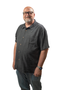 Portrait of a smiling, bald man with a gray beard and glasses wearing a dark button-up shirt.